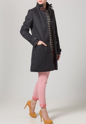 Vrouw draagt een donkergrijze jas met knopen over een gestreept shirt, roze opgerolde broek en mosterdgele hoge hakken, staand met één hand in de zak.