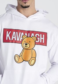 Sudadera blanca con un gráfico de un oso de dibujos animados en naranja y amarillo, con el texto "KAVANAGH" en rojo negrita encima del oso. Textura de tela suave.