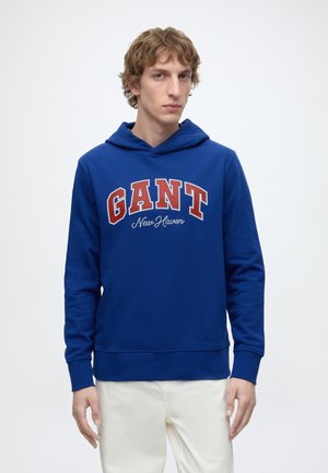 Junger Mann mit lockigem Haar, der einen blauen GANT New Haven Hoodie und weiße Hose vor einem einfarbigen weißen Hintergrund trägt.