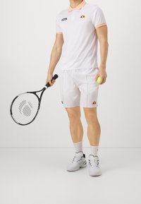 Homme en tenue de tennis blanche tenant une raquette noire dans sa main droite et une balle de tennis jaune dans sa main gauche, se tenant sur un fond uni.