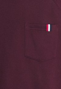 Poche en tissu bordeaux avec une étiquette rayée rouge, blanche et bleu marine cousue dans le coin supérieur droit de la poche.