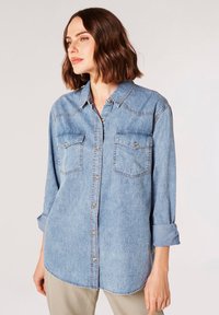 Blå denimskjorte med knaplukning fortil, krave, to brystlommer og opprullede ærmer. Har kontraststikninger og en afslappet pasform.