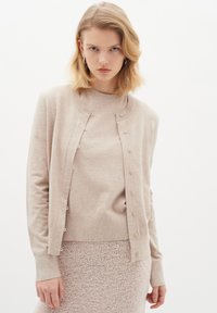 Hellbeige Strickjacke mit Knopfverschluss, Rundhalsausschnitt, langen Ärmeln und weicher Textur, getragen über einem passenden, strukturierten Strickrock.