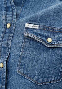 Blue denim shirt pocket with "Guess Jeans" label and pearl snap buttons, visible stitching on textured fabric.