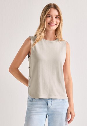 Junge Frau lächelt, trägt ein ärmelloses beiges Top mit seitlichen Knöpfen und hellblaue Jeans vor einfarbigem Hintergrund.