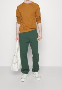 Sudadera marrón de cuello redondo, pantalones de chándal verdes con parche de logo, zapatillas blancas y un bolso de mano blanco texturizado. Atuendo casual con un ajuste relajado.