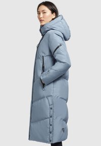 Donna che indossa un lungo cappotto invernale imbottito di colore azzurro chiaro, con cappuccio, tasche con zip e bottoni a pressione lungo la cucitura laterale.