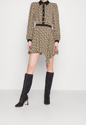 Femme portant une robe longue à manches longues beige et noire à motifs, avec un col noir et des boutons, associée à des bottes noires texturées montant jusqu'aux genoux.