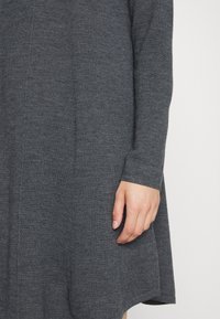 Gros plan sur la main d'une personne reposant contre un vêtement à manches longues en tricot gris foncé avec un détail de couture subtil.