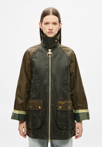 Σκούρο πράσινο και καφέ αδιάβροχο μπουφάν με φερμουάρ μπροστά, ψηλό γιακά, χρυσά μεταλλικά στοιχεία, δύο μπροστινές τσέπες και καρό λεπτομέρειες στα μανίκια.