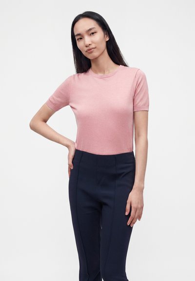 Jeune femme debout, main sur la hanche, portant un haut rose clair à manches courtes et un pantalon taille haute bleu marine foncé sur un fond uni.