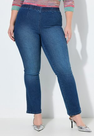 Plus-size person iført mørkeblå denim jeggings med elastisk talje og sølvfarvede spidse hæle.