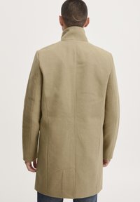 Cappotto in lana beige con colletto alto, taglio dritto e cuciture minime. Presenta tasche laterali e una texture pulita e liscia.