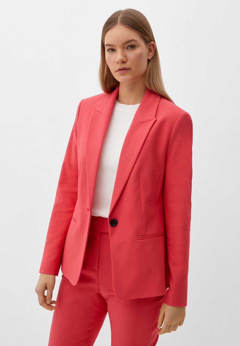 Blazer rose corail avec un bouton noir unique, revers cranté et deux poches avant. Assorti à un top blanc à col ras du cou. Texture lisse.