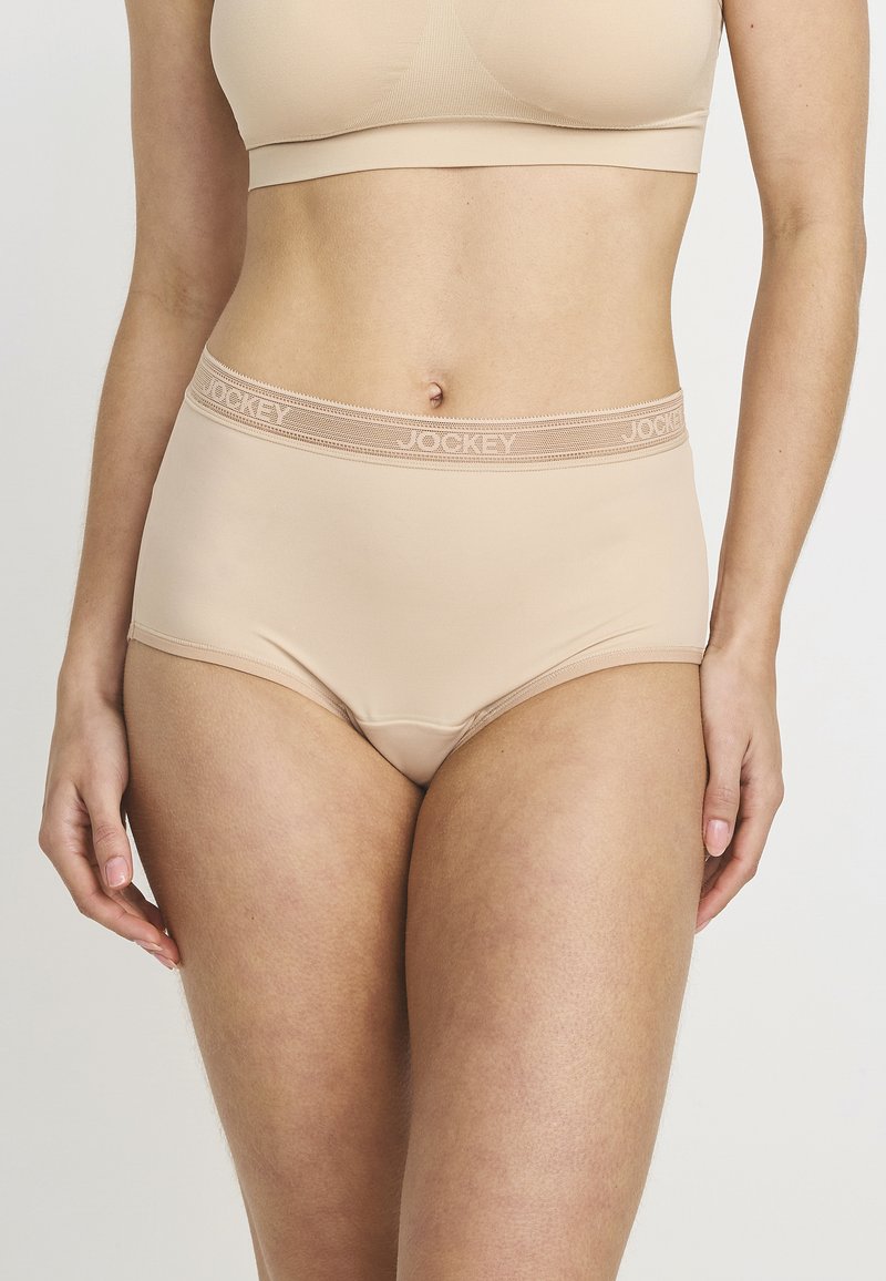 Vrouw in beige naadloze hoge taille onderbroek en bijpassende sportbh, die middenrif en bovenbenen toont tegen een effen achtergrond.