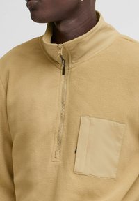 Fleece pullover in light beige with a half-zip, high collar, and a patch pocket on the front. Soft texture and smooth finish.