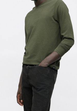 Personne portant une chemise olive à manches longues et un jean noir, une main dans la poche, sur un fond uni clair.