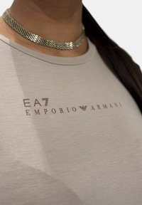 Camiseta ajustada de color gris claro con cuello redondo, con el logo impreso en color burdeos "EA7 EMPORIO ARMANI". Collar de cadena metálica visible.