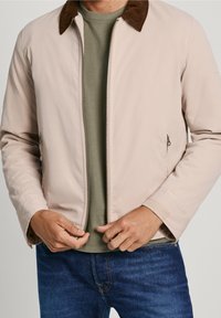 Lyserød bomberjakke med en brun fløjlskrave, lynlås foran og sidelommer, båret over en grøn t-shirt og blå jeans.