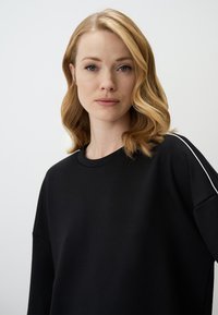 Zwarte sweatshirt met een ontspannen pasvorm, ronde halslijn en subtiele witte accenten langs de schouders. Zachte textuur, casual ontwerp.
