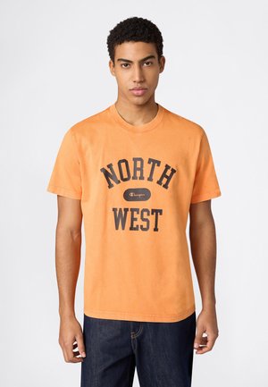 Młody mężczyzna w pomarańczowej koszulce z napisem "NORTH WEST" i logo Champion, w ciemnoniebieskich dżinsach, stojący na jednolitym tle.
