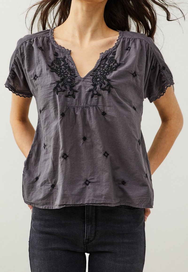 Blouse grise avec décolleté en V, manches courtes, ornée de broderies florales noires et de petits détails noirs. Tissu doux et texturé avec une coupe ample.