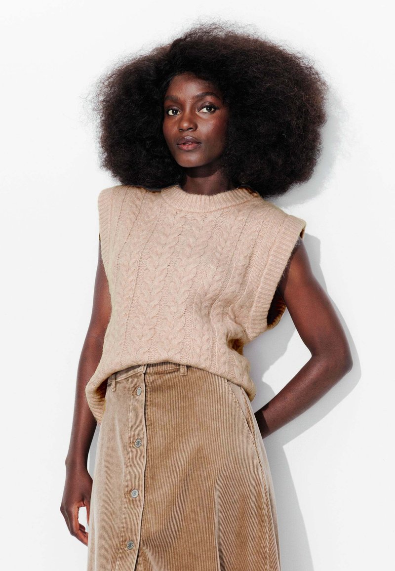 Pull sans manches en tricot cable beige avec un col côtelé, associé à une jupe en velours côtelé marron clair dotée d'une fermeture à boutons sur le devant.