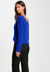 Maglione a maglia blu royale con scollo a V sul retro, maniche lunghe e polsini a costine. Abbinato a pantaloni neri, evidenziando il suo taglio rilassato.