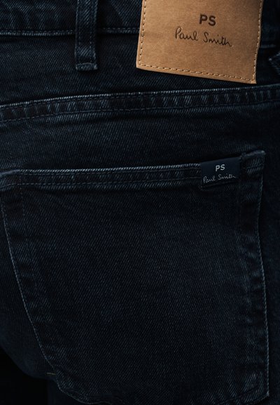 Tamsaus džinso audinio kelnės su odiniu ženkliuku, ant kurio parašyta „PS Paul Smith“. Audinys yra lygios tekstūros, su minimaliais išblukimais ir subtiliais siūlais.