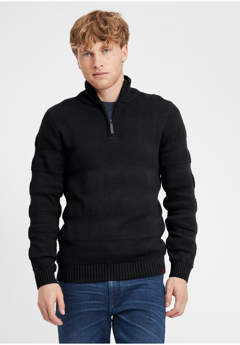 Maglione nero a maglia con colletto zip, caratterizzato da un motivo a strisce testurizzato. Polsini e orlo a coste, indossato su jeans blu.