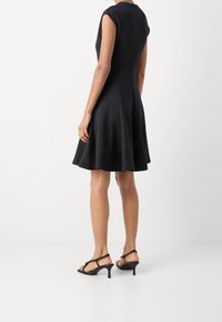 Robe noire avec un corsage ajusté et une jupe évasée, manches courtes style trapèze et fermeture éclair cachée dans le dos. Portée avec des sandales noires à talons.