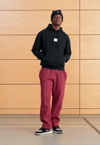 Zwarte hoodie met een voorlabel, bordeauxrode joggingbroek en zwarte sneakers met witte accenten. Op de achtergrond lichte houten panelen.