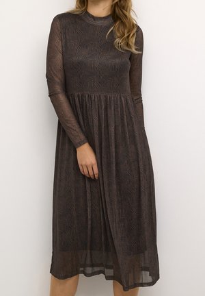 Frau trägt ein transparentes, dunkelbraunes Midi-Kleid mit langen Ärmeln und abstrakten geometrischen Mustern und steht vor einem einfarbigen hellen Hintergrund.