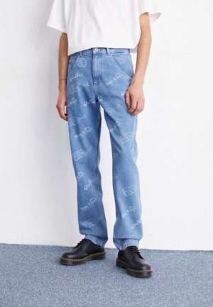 Jeans de color azul claro con un corte recto, que presentan patrones impresos en blanco. Combinados con zapatos negros de cordones y una camisa blanca de manga corta.