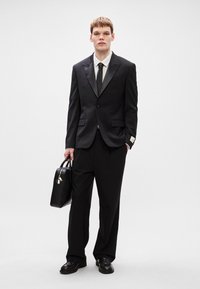 Μαύρο κοστούμι με σακάκι ενός κουμπιού, συνδυασμένο με πτυχωτό παντελόνι. Λευκό πουκάμισο με ρίγες, μαύρη γραβάτα και μαύρα παπούτσια. Φορώντας μια μαύρη τσάντα.