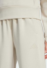 Pantalones de chándal beige de Adidas hechos de una tela suave, con una cinturilla elástica, bolsillos laterales y un logotipo elevado en la parte cerca de la rodilla.