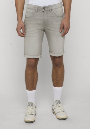 Mężczyzna ubrany w jasnoszare dżinsowe szorty z podwiniętymi brzegami, białe skarpetki i beżowe buty New Balance, stojący na tle jednolitego tła.