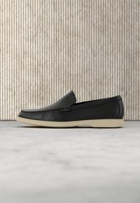 Mocassin en cuir noir à enfiler avec dessus texturé et semelle en caoutchouc beige sur fond gris texturé.