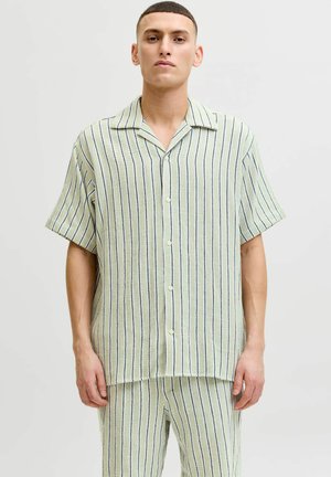 Homme portant une chemise à manches courtes boutonnée vert clair et un pantalon assorti avec de fines rayures verticales noires et blanches, debout devant un fond uni.