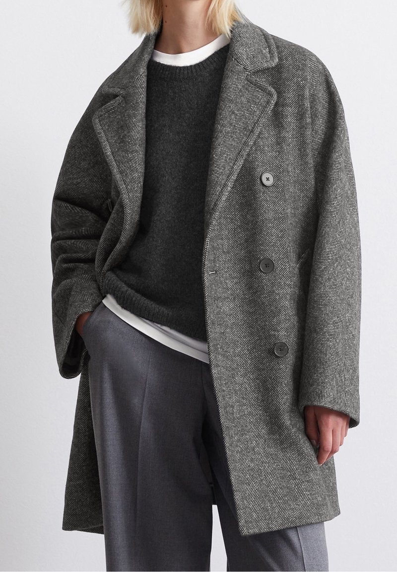 Personne portant un manteau croisé à chevrons gris, pull gris foncé, chemise blanche en dessous, et pantalon gris, debout avec une main dans la poche.