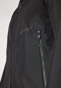 Veste légère noire avec une fermeture éclair frontale complète et une poche latérale zippée. Le tissu a une texture lisse, avec un logo imprimé sur la poitrine.