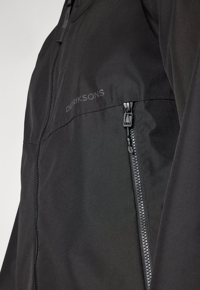 Schwarze leichte Jacke mit einem durchgehenden Reißverschluss vorne und seitlichen Taschen mit Reißverschluss. Der Stoff hat eine glatte Textur, mit einem Logo auf der Brust bedruckt.