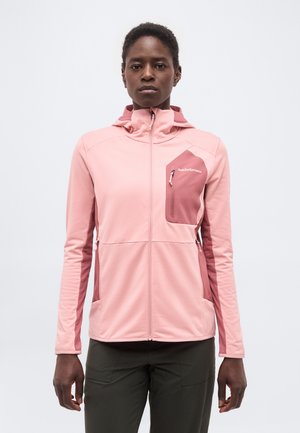 Kobieta w różowej bluzie z kapturem zapinanej na zamek, z kieszenią na piersi i kontrastowymi panelami bocznymi, stojąca na jednolitym tle.