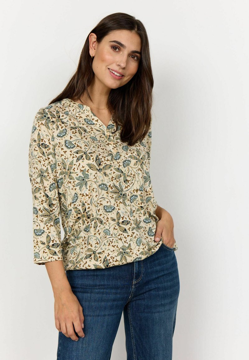 Bluse med blomstermotiv på lys baggrund, blå og grønne blomster, trekvartærmer og en lille knaplukning. Kombineret med blå jeans.