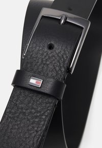 Ceinture en cuir noir avec une finition texturée, boucle en métal argenté et un petit emblème featuring des rectangles rouges, blancs et bleus.
