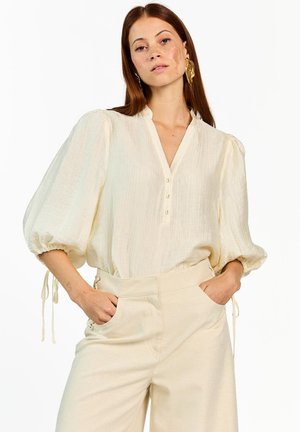 Vrouw met lang rood haar, gekleed in een crème textuur blouse met pofmouwen en beige hoge taille broek, handen in de zakken, neutrale achtergrond.