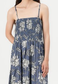 Blaues Blumendruckkleid mit dünnen Trägern, gerafftem Oberteil und gestuftem Rock. Verziert mit weißen Blumenmustern auf strukturiertem Stoff.