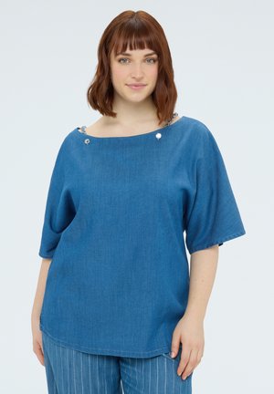 Žena s krátkými hnědými vlasy v modrém volném topu se stříbrnými knoflíky na ramenou a modrých kalhotách s proužky, stojící na bílém pozadí.