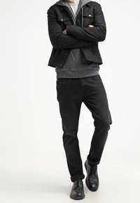 Veste noire avec poches à boutons sur un sweat à capuche gris, associée à un jean noir et des chaussures noires. Le mannequin est debout, les bras croisés.