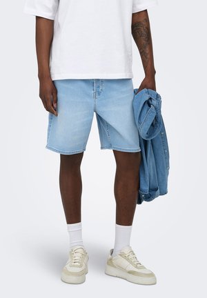 Personne portant un short en jean bleu clair, des chaussettes blanches, des baskets blanches, un t-shirt blanc et tenant une veste en jean bleue dans une main.